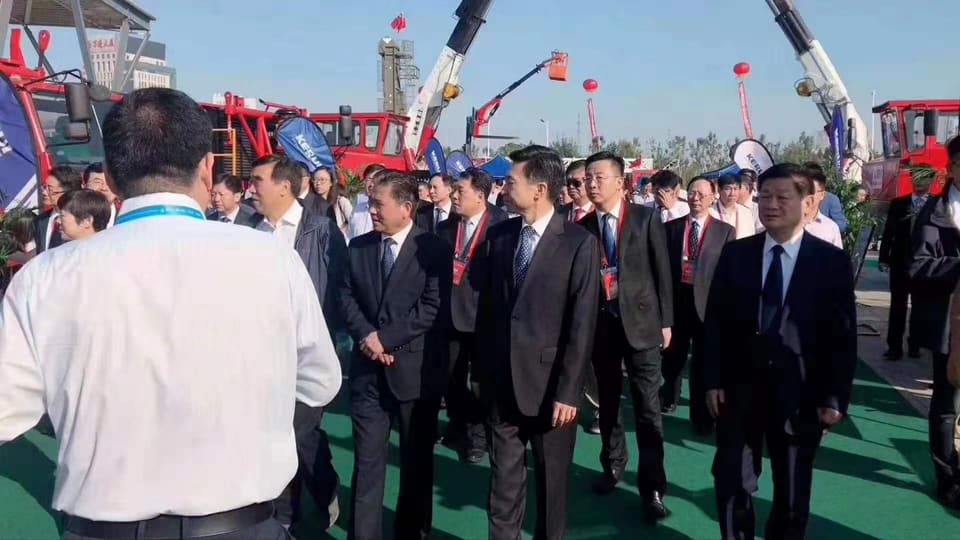 Visitors in suits walk past cranes and heavy machinery on the outdoor exhibition grounds.